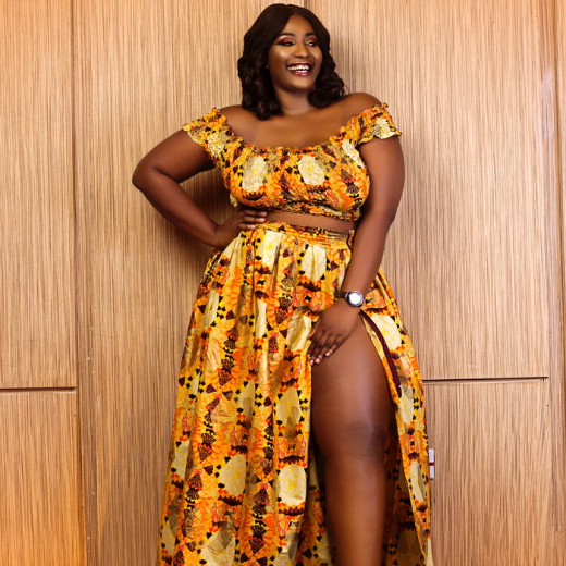 A woman wearing co-ord off shoulder top and high slit skirt in African print against a brown background.